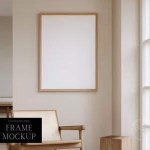 May include: A wooden frame mockup with a white background, hanging on a light beige wall. The frame is in a living room setting with a wooden chair and a wooden stool.