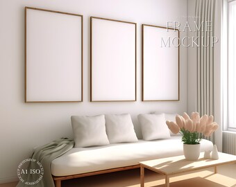 Op de afbeelding: Drie lege fotolijsten met houten frames hangen aan een witte muur boven een witte bank met kussens. Een vaas met roze bloemen staat op een houten salontafel voor de bank.