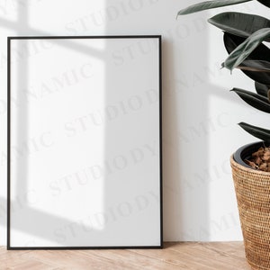May include: A black picture frame with a white mat and a white background. The frame is sitting on a light wood floor. A potted plant with dark green leaves is in the background.