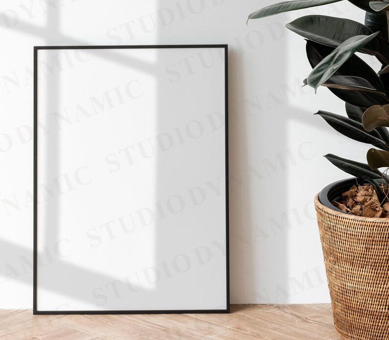 May include: A black picture frame with a white mat and a white background. The frame is sitting on a light wood floor. A potted plant with dark green leaves is in the background.