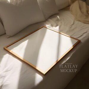 May include: A gold picture frame with a white mount on a white bed with a white pillow and a beige throw. The text "FLATLAY MOCKUP" is in the bottom right corner of the image.
