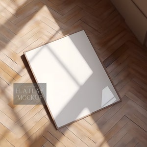 Op de afbeelding: Een witte frame mockup op een houten vloer met een visgraatpatroon. De frame is schuin geplaatst, waardoor schaduwen op de vloer vallen. De tekst "FLATLAY MOCKUP" is zichtbaar in de linkerbenedenhoek van de afbeelding.