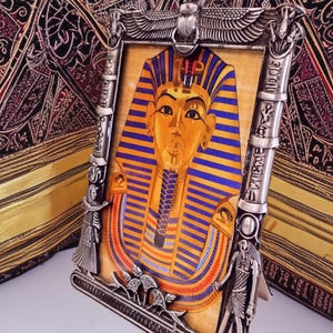 May include: Silver-toned picture frame with an Egyptian theme. The frame surrounds an image of King Tutankhamun. The frame has detailed carvings of Egyptian figures and symbols, including a scarab beetle at the top. The image features gold, blue, and red colors.