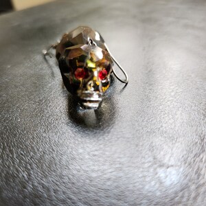 May include: A crystal skull earring with red gemstone eyes. The earring is silver-toned and has a dangling design.