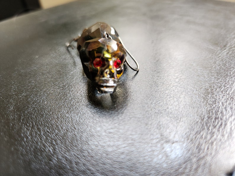 May include: A crystal skull earring with red gemstone eyes. The earring is silver-toned and has a dangling design.