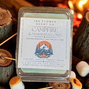 May include: A square, clear container of soy wax melts with the scent "Campfire & Marshmallows" by Ink Flower Scent Co. The label includes a mountain and tree graphic. The wax melts are 2.5 oz (70.8g). Marshmallows on sticks and wood slices are in the foreground.