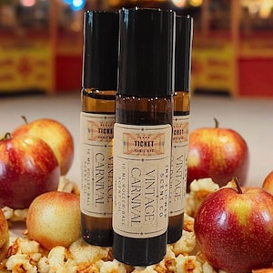 May include: Three rollerball scent bottles with black caps and labels that read "Vintage Carnival." The bottles are surrounded by red apples and popcorn. The labels also read "Ink Flower Scent Co."