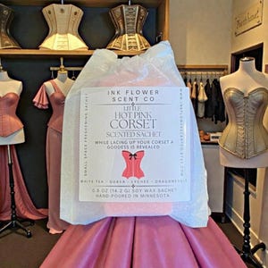 May include: A pink scented sachet labeled "Little Hot Pink Corset" by Ink Flower Scent Co. The sachet is in a clear bag, with a pink corset graphic. The sachet is 0.5 oz (14.2 g) and hand-poured in Minnesota.