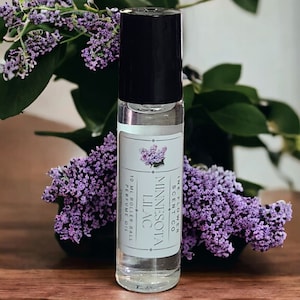 May include: A clear glass rollerball bottle with a black cap and a white label. The label reads "Minnesota Scent Co. Ink Flower Lilac 10 ml Roller-Ball Perfume Oil". The bottle is sitting on a wooden surface next to a sprig of purple lilac flowers.
