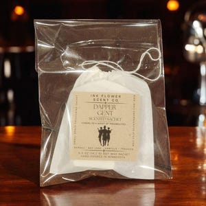 May include: A small, clear plastic bag containing a white sachet with the text "Dapper Gent Scented Sachet." The sachet has a beige label with the words "Ink Flower Scent Co." and a list of scents. The sachet is hand-poured in Minnesota and weighs 0.5 oz.