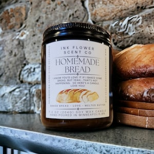 May include: A brown glass candle jar with a white label that reads "Ink Flower Scent Co. Homemade Bread". The label features a watercolor illustration of a loaf of bread and the text "I know you'd love it if I baked some bread, but yeah... that's not happening. So here's a candle. Love you!" The label also includes the text "Baked Bread - Love - Melted Butter" and "7.2 oz (204g) Soy Wax Candle Hand-Poured in Minneapolis, MN".
