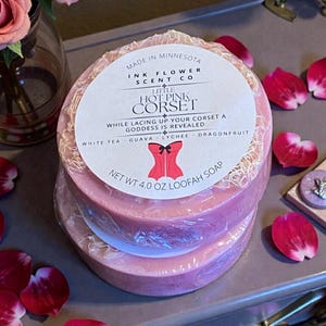May include: Pink loofah soap bars, stacked and wrapped in clear plastic. The soap is round with a light pink hue and a loofah center. The label reads "Little Hot Pink Corset" with scent notes. The net weight is 4.0 oz.