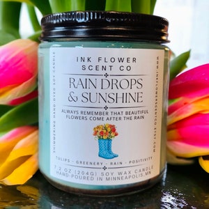 May include: A 7.2 ounce soy wax candle in a glass jar with a black lid. The label is white with blue text and a floral illustration. The candle is called "Rain Drops & Sunshine" and is hand-poured in Minneapolis, Minnesota.
