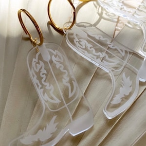 May include: Clear acrylic cowboy boot earrings with gold-colored hoops. The boots are engraved with a white design and the text "Clear & White Engraved" is visible.