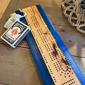 Op de afbeelding: Een houten cribbagebord met een blauwe basis en een pakje Bicycle speelkaarten. Het cribbagebord is gemaakt van lichtgekleurd hout met gelijkmatig verdeelde gaten voor de pinnen. Er is ook een klein beige zakje met trekkoord aanwezig.