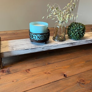 May include: Whitewashed wooden tray with brown wood accents, featuring a blue candle in a metal holder, a glass vase with dried oats, and a green topiary ball.