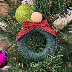 以下が含まれることがあります： 手作りのクリスマスオーナメント。 濃い緑色の小さなリースに紐が巻かれ、赤と黒のチェック柄のリボンと木製のビーズが付いています。 オーナメントはクリスマスツリーに飾られています。
