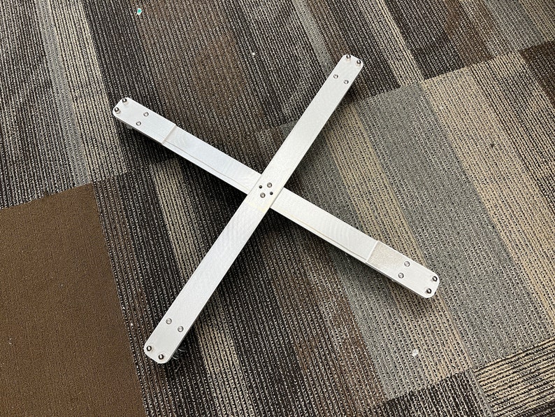 May include: A silver metal cross-shaped base with four arms and multiple holes. The base is lying flat on a patterned carpet.