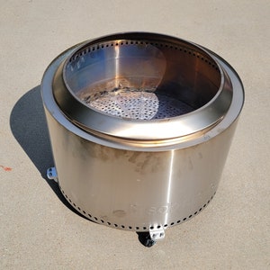 May include: A stainless steel fire pit with a round bowl and a perforated base. The fire pit has a brushed finish and is designed for outdoor use.