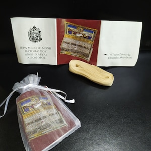 Puede incluir: Una pequeña bolsa de organza blanca con un libro rojo, dorado y blanco en su interior. El libro tiene una imagen de una escena religiosa en la portada. La bolsa está sobre una superficie negra junto a una cinta blanca y un folleto doblado con una imagen de una caja dorada y plateada con una escena religiosa. El folleto tiene texto en griego que dice "ΙΕΡΑ ΜΕΓΙΣΤΗ ΜΟΝΗ ΒΑΤΟΠΑΙΔΙΟΥ 630 86 ΚΑΡΥΑΙ ΑΓΙΟΝ ΟΡΟΣ" y "Η Τιμία Ζώνη της Υπεραγίας Θεοτόκου".