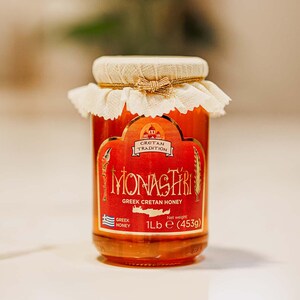 May include: A glass jar of honey with a red label that says "Monastiri Greek Cretan Honey" and a white label that says "Cretan Tradition". The jar is tied with a white ribbon and has a gold lid. The jar is sitting on a white surface.