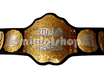 May include: A gold and black wrestling championship belt with the text "TNA WRESTLING HEAVYWEIGHT CHAMPION". The belt has gold accents and red jewels. The belt is made of black leather.