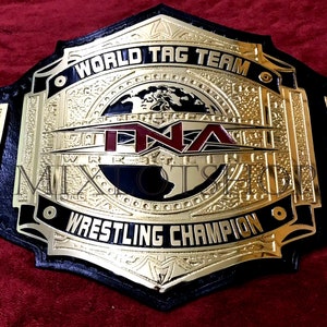 May include: A gold and black wrestling championship belt with the text "WORLD TAG TEAM" and "WRESTLING CHAMPION". The belt features the letters "TNA" in red. The belt is made of leather and gold-colored metal.