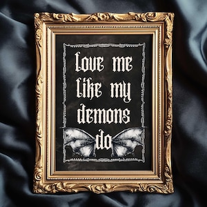 Puede incluir: Una impresión enmarcada con el texto "Love me like my demons do" en letras góticas. La impresión presenta un fondo negro con alas de murciélago blancas en la parte inferior. El marco es ornamentado y dorado, sobre una tela azul oscuro.