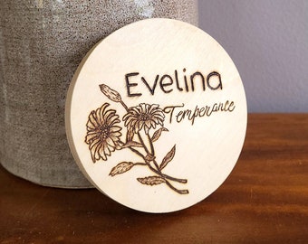 May include: A round wooden sign with the name "Evelina Temperance" burned into the surface. The sign is decorated with a floral design of three flowers.