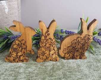 May include: Three wooden bunny figurines with floral designs carved into them. The bunnies are standing on a green fabric surface.