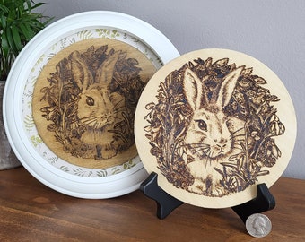 May include: Two wood-burned round plaques with a white rabbit in a field of flowers. The smaller plaque is on a black stand. The larger plaque is in a white frame.