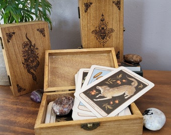 May include: Three wooden boxes with engraved designs. The boxes are closed, except for one which is open and contains a deck of cards with illustrations of animals and plants. The text on one card reads "The Wolf and Rose Hip".