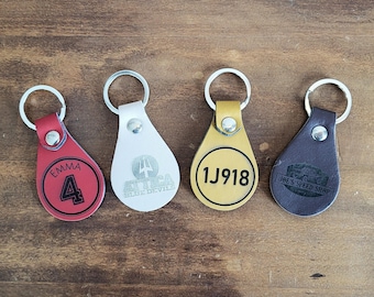 May include: Four leather keychains with different designs. One keychain is red with the name Emma and the number 4. Another keychain is white with the text "STICA BLUE DEVILS" and a trident symbol. A third keychain is yellow with the number "1J918". The final keychain is brown with the text "JOE'S SPEED SHOP" and a car graphic.