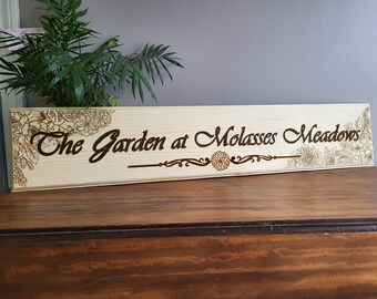 May include: A wooden sign with the text "The Garden at Molasses Meadows" burned into the surface. The sign is decorated with floral designs.