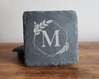 May include: A set of four gray slate coasters with a geometric design and a single initial "M" in a white font. The coasters are decorated with a leafy design.