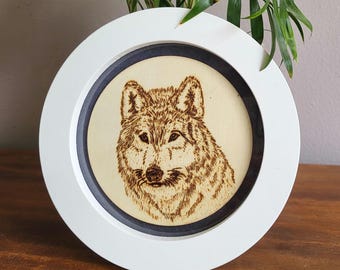 May include: A white wood frame with a black inner border surrounds a wood burned image of a wolf's face.