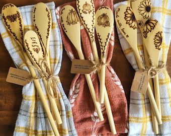 May include: Six wooden spoons with various designs and sayings are tied together with twine and displayed on two kitchen towels. The spoons feature designs of sunflowers, pumpkins, bees, and the sayings "Give me some honey", "Made with love", "Hello Fall", and "Spread with love".