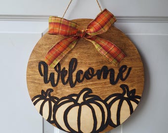 May include: A round wooden door hanger with a welcome sign and three pumpkins. The door hanger is decorated with a plaid bow in orange, red, and yellow.