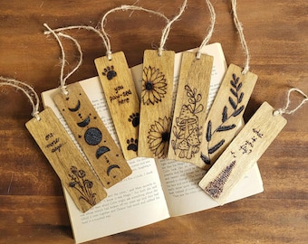 May include: Eight wooden bookmarks with different designs, including a paw print, a sunflower, mushrooms, a tree, and a moon phase. The bookmarks are tied with twine and have a natural wood finish.