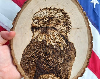 May include: A wood-burned image of a bald eagle on a round wooden plaque. The eagle is depicted in detail with brown and tan tones, set against a backdrop of the American flag. The plaque has a natural, rough bark edge.