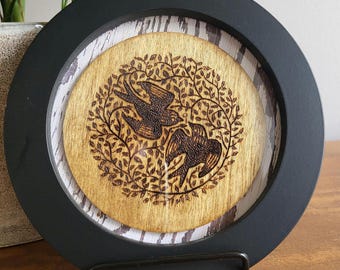 May include: A black framed wood wall decor with a burnt wood design of two birds in a circle of leaves.