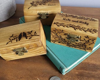 May include: Three small wooden boxes with engraved designs and names. The boxes have a hinged lid and a brass latch. The boxes are sitting on a book and a wooden surface.