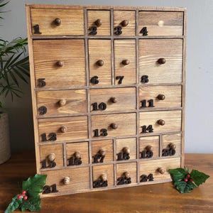 Può includere: Un calendario dell'Avvento in legno con 24 cassetti numerati. I cassetti sono disposti a griglia e hanno piccole manopole rotonde. Il legno ha una calda tonalità marrone. I numeri sono in un carattere nero perlato.