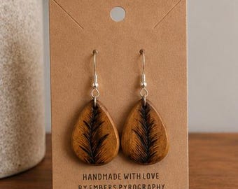 May include: A pair of teardrop-shaped wooden earrings with a dark, feather-like design. The earrings are attached to silver hooks and displayed on a brown card. The card reads "HANDMADE WITH LOVE BY EMBERS PYROGRAPHY Sterling Silver."
