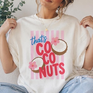 May include: Cream-colored t-shirt with the text "that's COCO NUTS" in blue and red, featuring coconut illustrations with pink flowers. The shirt has a pink striped background.