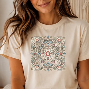 May include: A cream-colored t-shirt featuring a square, symmetrical design in shades of red, blue, green, and beige. The design has floral and geometric elements. The shirt is short-sleeved and made of a soft material.
