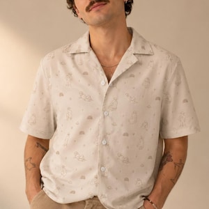 May include: A short-sleeved button-down shirt in a light beige color featuring a repeating pattern of thin, brown line-art illustrations of figures and small rainbows. The shirt has a classic collar, a front button placket, and a relaxed fit.