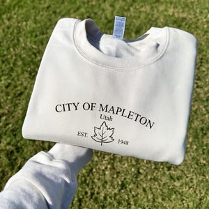 Könnte beinhalten: Cremefarbenes Sweatshirt mit dem Text "CITY OF MAPLETON Utah" und einer Blattgrafik. Unter dem Blatt steht der Text "EST. 1948". Das Sweatshirt ist gefaltet.