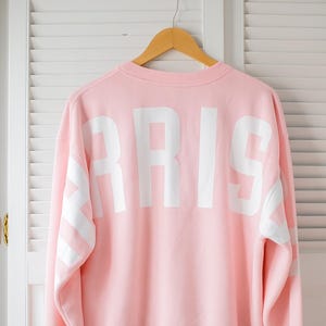 Puede incluir: Una sudadera rosa claro con la palabra "PARIS" impresa en letras blancas grandes en la espalda. Las mangas tienen rayas blancas. La sudadera está colgada en una percha de madera.