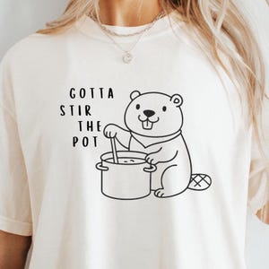 Puede incluir: Camiseta color crema con un castor de dibujos animados removiendo una olla, con el texto "GOTTA STIR THE POT" en negro. La ilustración del castor es un contorno simple, dando un toque juguetón al diseño.